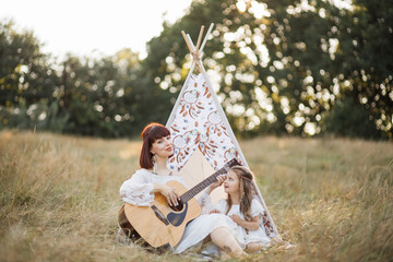 Charming young boho hippie woman playing guitar to her cute little daughter outdoor in nature on sunny evening, sitting in front of wigwam tent and singing. Musical family traditions © Sofiia Shunkina