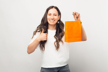 Positive young woman is showing like gesture and a shopping bag over white background.