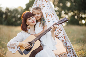 Pretty hippie woman sits on the grass near teepee tent outdoors, having fun playing guitar for her...
