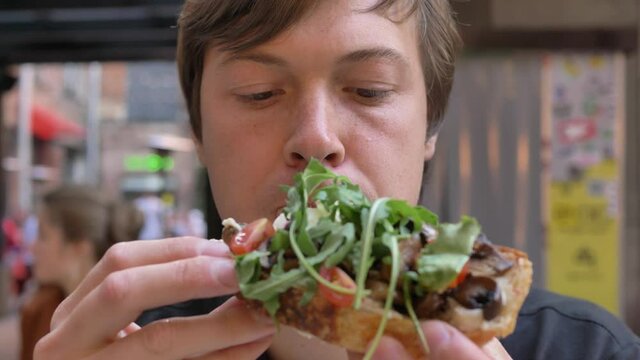 Young man holds in hands and eats with mouth full canape with mushrooms ruccola and tomatoes for breakfast in cozy cafe closeup