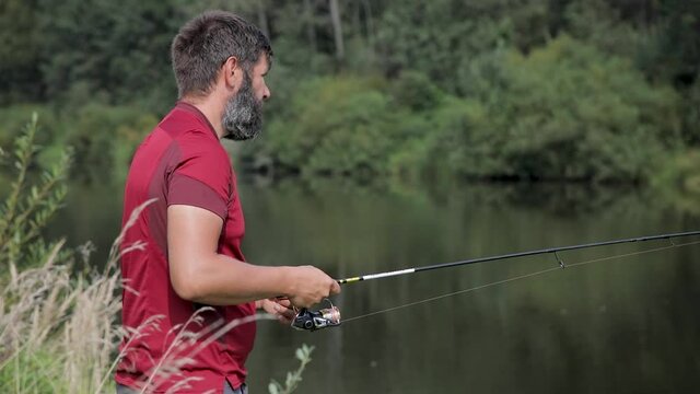 A man with a beard is fishing on the river, casting a line. A fisherman with a fishing rod is fishing on the river bank.