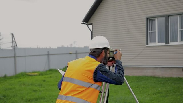 A male engineer works with the optical level on site. A builder in a helmet looks into an optical device.