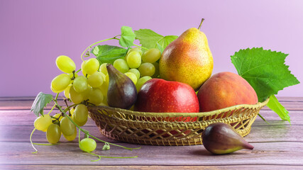 Juicy, bright, ripe fruits in a wicker basket on a pink background stand on a wooden table. Bunch of grapes, grapevine, red apple, yellow pear and figs.