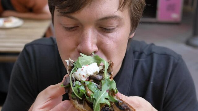 Young man holds in hands sandwich with mushrooms and ruccola and eats biting over wooden table in cozy cafe at breakfast closeup