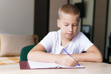 My favorite subject. Handsome smiling blonde boy is working and writing in his notebook while sitting at table, doing his homework on the school subject. Caucasian boy writes in a notebook with a pen