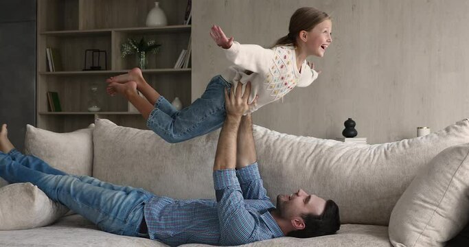 Young strong father lying on couch lift up on outstretched hands little cute daughter, 8s child imitates plane imagines herself like flying in air. Funny leisure activities on weekend at home concept