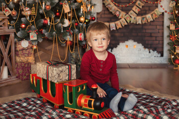 Obraz na płótnie Canvas Portrait of happy child 2-3 years old sits in New Year's decorations at home under a decorated Christmas tree and waits for gifts. Alone baby boy playing in train toy.