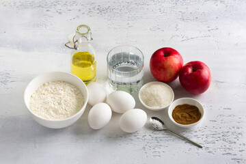 Ingredients for homemade apple pie: apples, flour, eggs, water, oil, sugar, cinnamon, salt on a light gray textured background, top view