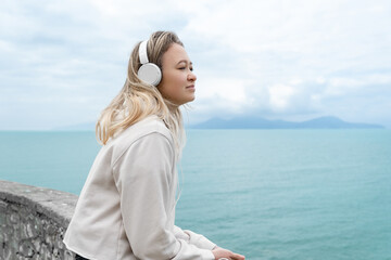 Fototapeta na wymiar Asian Young Girl Listening Track or Podcast With White Over-Ear Headphone On The Mediterranean Sea. Leisure Meditation for Calming Down and Relaxing. Mindfulness and Music Therapy.