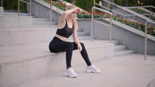 Fitness young woman wipes sweat sitting stairs park after hard morning workout training. Caucasian athletic female resting after exercises outdoors dressed black sportswear Blonde hair 25 years old 