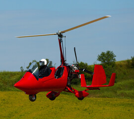 Fototapeta na wymiar Red open-cockpit gyroplane landing over green field on sunny day