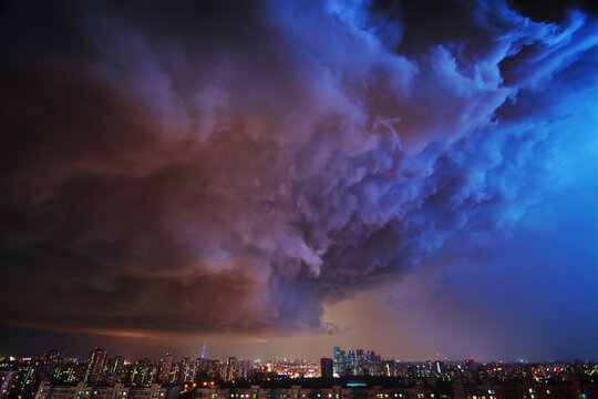 Amazing view of the night storm over the big city, with colorful clouds moving on the sky and bringing first drops of rain, and bright lightnings illuminating the scene.