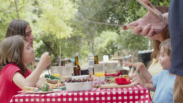 Slider shot of happy family eating delicious food at picnic. Caucasian father putting freshly cooked kebab on plate. Barbeque, family dinner, spending time together concept
