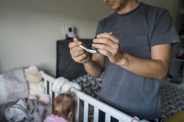 Father preparing spoon with probiotic drops medicine for his small caucasian baby girl while standing by the cradle crib at home