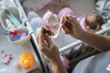 Father preparing spoon with probiotic drops medicine for his small caucasian baby girl while standing by the cradle crib at home close up on hands selective focus copy space care and parenting concept