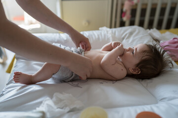 Obraz na płótnie Canvas Side view on caucasian baby lying on the bed while her mother is changing diapers and clothes four months old daughter at home - parenting childhood new life and growing up concept