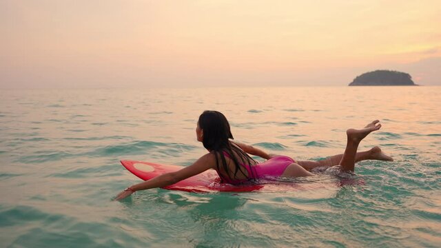 4K Confidence Asian woman floating on surf board passing beautiful sea and twilight sky at summer sunset. Female enjoy outdoor lifestyle activity and extreme water sport on summer holiday vacation