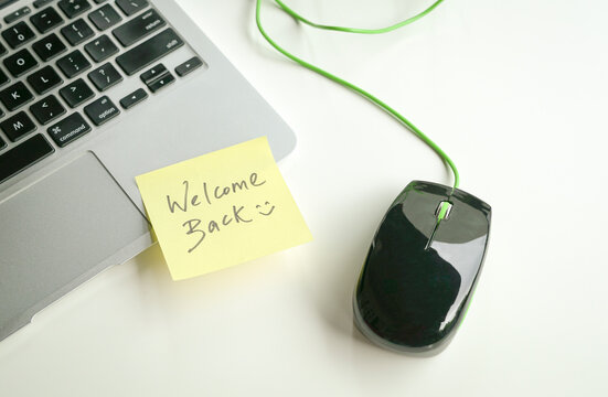 Welcome back with smiley written on yellow paper stick on keyboard next to computer mouse.
