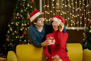 Man blindfold his lover and hold the present to make surprise to her during Christmas festival and they celebrate at home with fairly light and decorating as background.