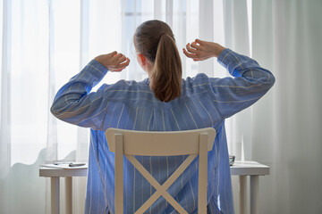 Back view of young female sitting at desk and stretching. Remote work challenges and negative...
