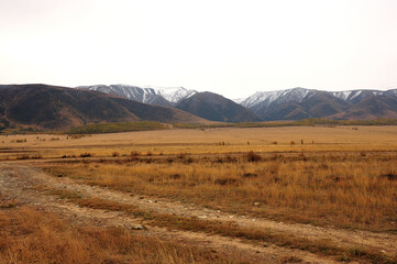 Fototapeta premium A field road going through the autumn steppe at the foot of a mountain range with snow-capped peaks.