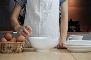 Close-up front view footage, a female cook in a white apron is cracking an egg into a cup to prepare a meal on a wooden table in the home's kitchen. Eating egg yolks is a healthy breakfast.