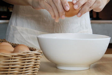 Close-up front view footage, a female cook in a white apron is cracking an egg into a cup to prepare a meal on a wooden table in the home's kitchen. Eating egg yolks is a healthy breakfast.