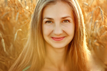 A large portrait of a young woman in a wheat field at sunset. The woman looks at the camera with a smile.