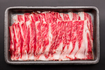 Sliced raw beef place in a row on a black plastic tray isolated on a black background. Sliced ​​meat for cooking, fresh meat for grilling, yakiniku, sukiyaki or shabu.