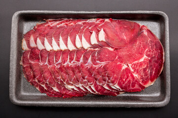 Sliced raw beef place in a row on a black plastic tray isolated on a black background. Sliced ​​meat for cooking, fresh meat for grilling, yakiniku, sukiyaki or shabu.