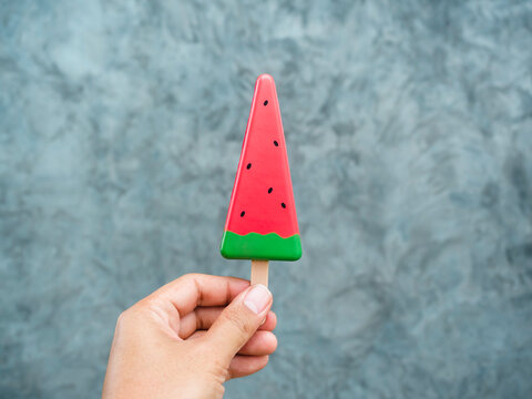 Hand holding artificial watermelon slice popsicle on grey concrete wall background. Summer fruit ice cream stick.