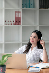 Portrait of young beautiful asian woman holding pen, smiling and looking outside while sitting in modern office room.