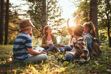 Kids raising hands asking questions during outdoor lesson with teacher in forest on sunny spring day