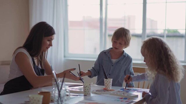 Group of kids and their teacher leaning over table with watercolor paints and painting self-made clay items. Children with teacher in art class.