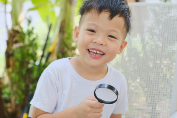 Cute happy smiling little Asian  5 years old kindergarten boy child exploring environment by looking through a magnifying glass in sunny day in garden, kid first experience and discovery concept