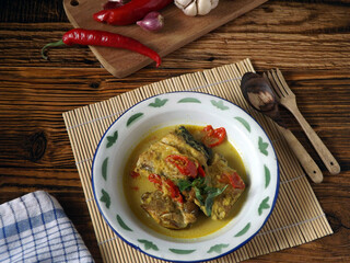 Mangut pe or spicy fish curry is Javanese cuisine. Smoked stingray fish cooked with spices and coconut milk. Served in enamel plate with ingredients around. Focus selection, rustic wooden background. 