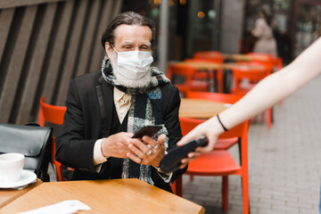 White senior man in face mask paying with mobile phone at cafe