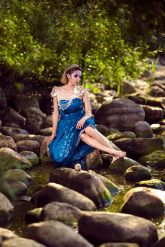 Relaxed Positive Lady Posing As Sea Mermaid  in Artistic Blue Dress And Strasses on Face Sitting in Wet Dress on Rocky Shore Outdoor.