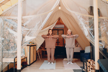 Family doing exercises sports indoors. Young and senior elderly woman relaxing at glamping camping tent. Mother and daughter modern vacation lifestyle concept