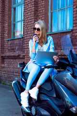 Naklejka premium Caucasian Female Biker in Blue Suit Working With Laptop While Sitting on Shiny New Motorcycle With Cup of Coffee Outdoors.