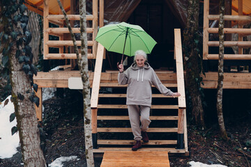Elderly mature woman with umbrella at glamping camping tent. Modern vacation lifestyle concept