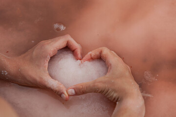 Women's heart-shaped hands hold pink salt on water surface of unique pink lake. Original color of lake is given by halophilic microalgae Dunaliella salina. Salt used in cosmetology and folk medicine