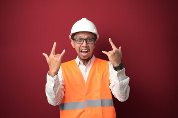 Young Asian worker man wearing helmet with crazy expression doing rock symbol with hands up