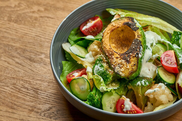 Diet salad with grilled avocado, white sauce and vegetables in a bowl on a wooden background with noise grain