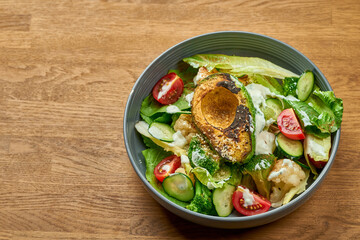 Diet salad with grilled avocado, white sauce and vegetables in a bowl on a wooden background with noise grain
