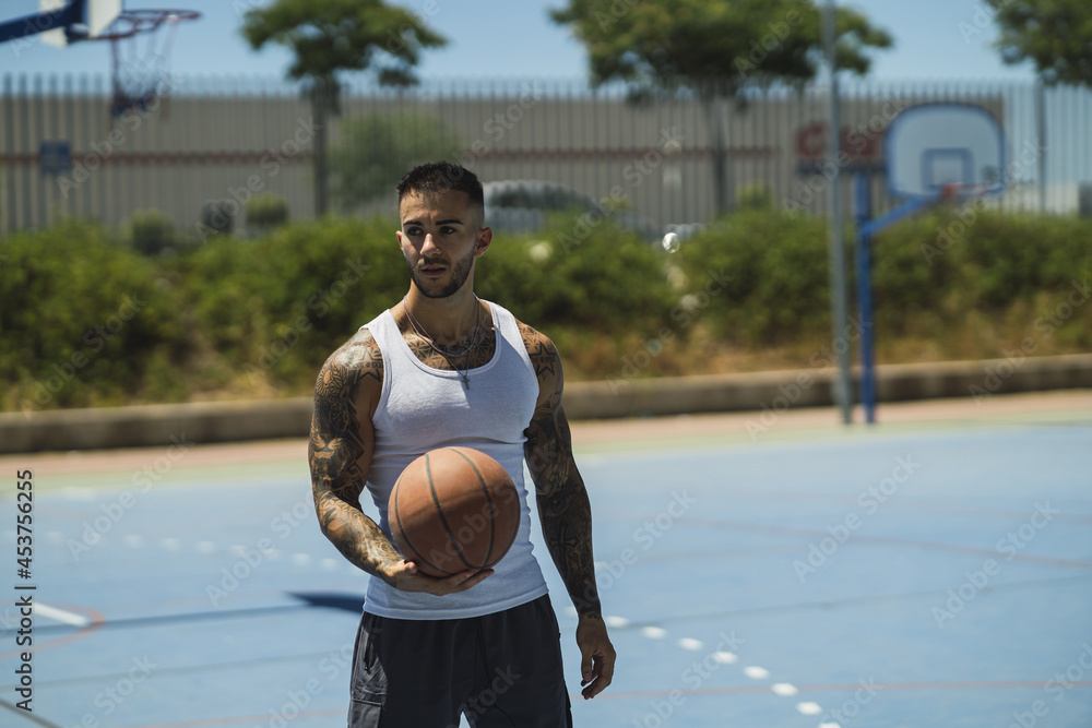 Wall mural handsome caucasian basketball player with cool tattoos standing on the court holding the ball