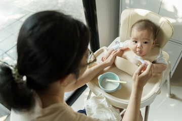 Young Asian Mother feeding to her Adorable baby boy with porridge or pureed food in high seat at home