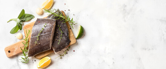 Top view of two raw halibut fish steaks with herbs and lemon on wooden board and white background....