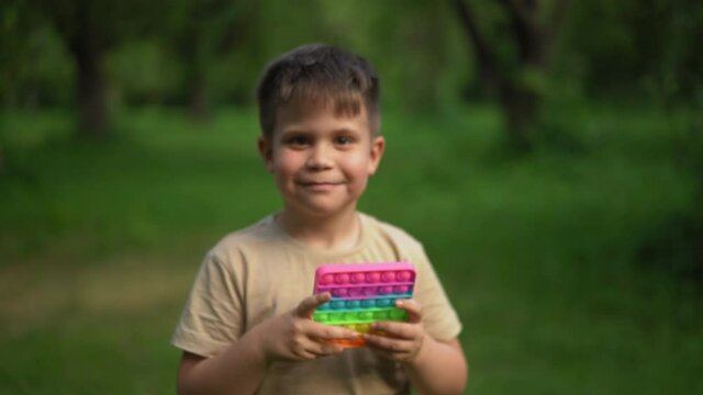 Child Smiles With an Antistress Toy and Takes Step Back. Slow Motion Video of Child Walking Away with toy in Hand. Kid Holding Rainbow Pop It Fidget Toy. High quality 4k footage