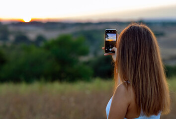 A young girl from behind with long blond hair, dressed in white, takes pictures of setting sun with...
